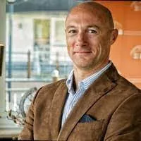 Foto do rosto de um homem branco, calvo, de terno marrom